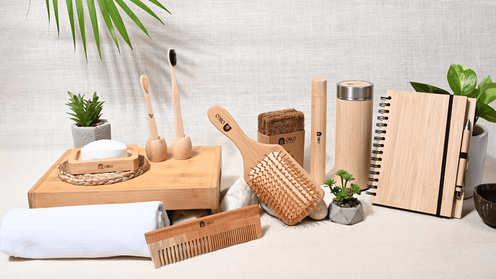 Collection of bamboo products including brushes, a comb, and containers on a light surface with plants in the background By oiko.com